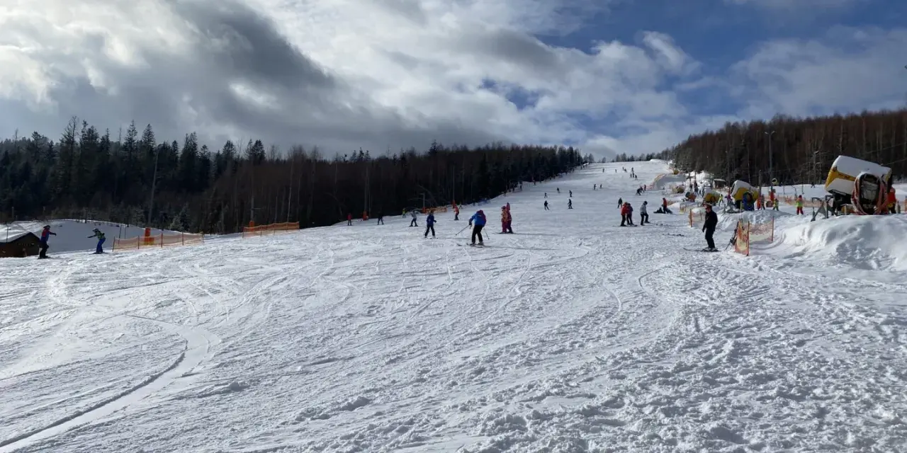 Słoneczny dzień na otwartych stokach narciarskich Wisła. Narciarze i snowboardziści cieszą się jazdą po ośnieżonych trasach, a w tle widać las i maszyny do naśnieżania.