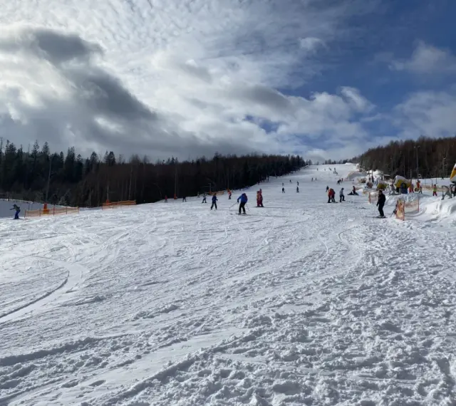 Słoneczny dzień na otwartych stokach narciarskich Wisła. Narciarze i snowboardziści cieszą się jazdą po ośnieżonych trasach, a w tle widać las i maszyny do naśnieżania.