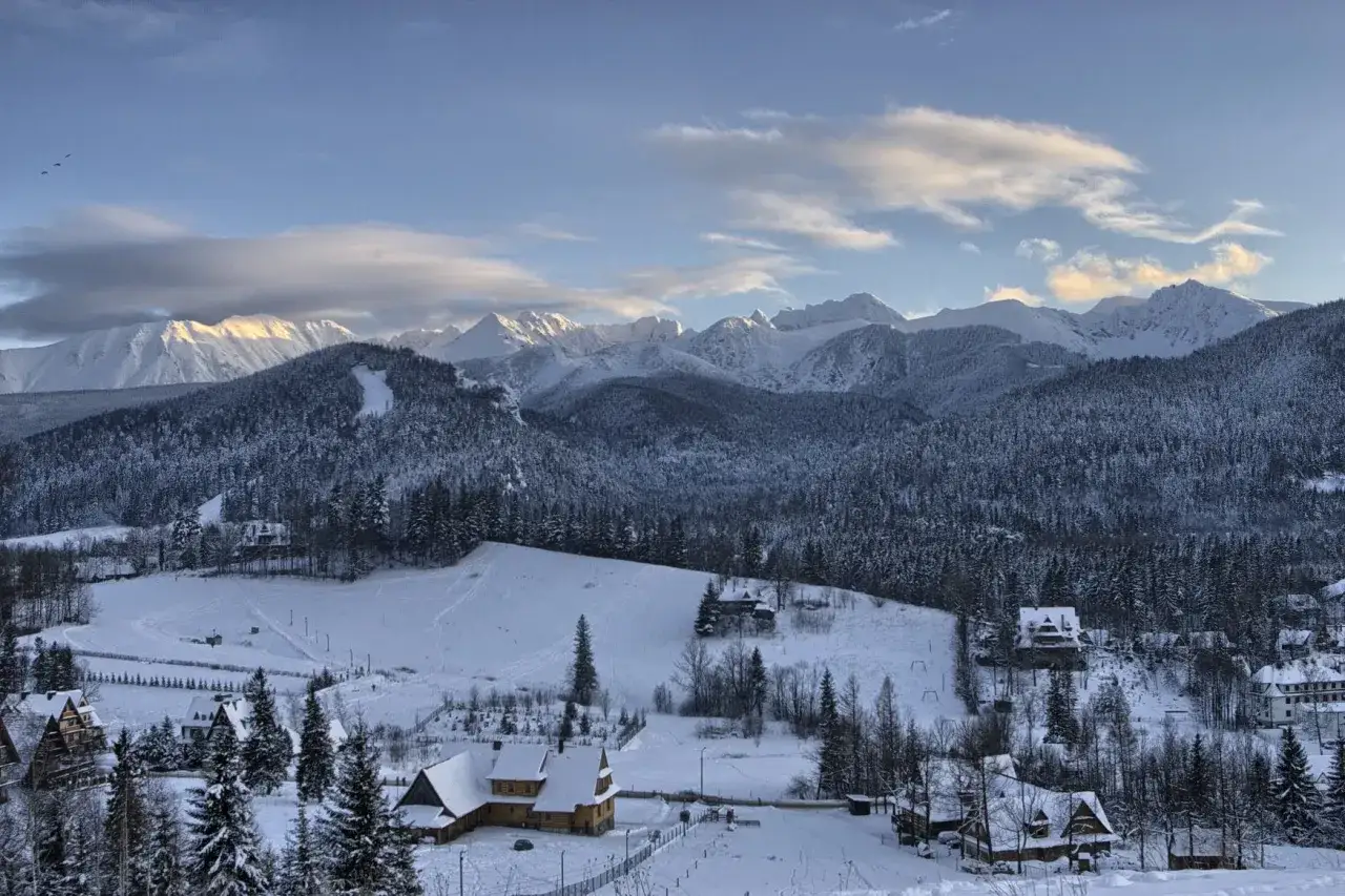 Zimowy krajobraz z górami w tle, pokryty śniegiem las i domki. Widać stoki narciarskie Zakopane.